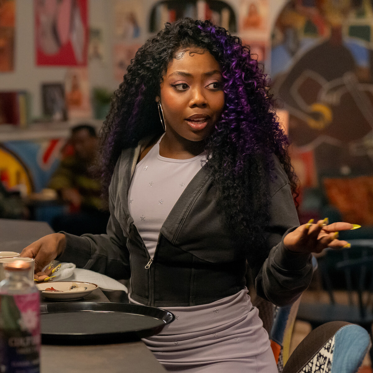 A woman with long curly hair and purple highlights sits at a table in a vibrant, art-filled café, gesturing with her hand while holding a tray and partially eaten food, with colorful artwork visible on the walls behind her.