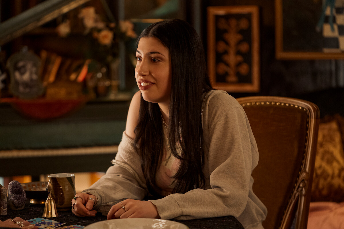 Young woman with long dark hair sits at a table with tarot cards, crystals, and a mug in a warmly lit, cozy room decorated with paintings, books, and vintage furniture, suggesting a mystical, relaxed atmosphere.