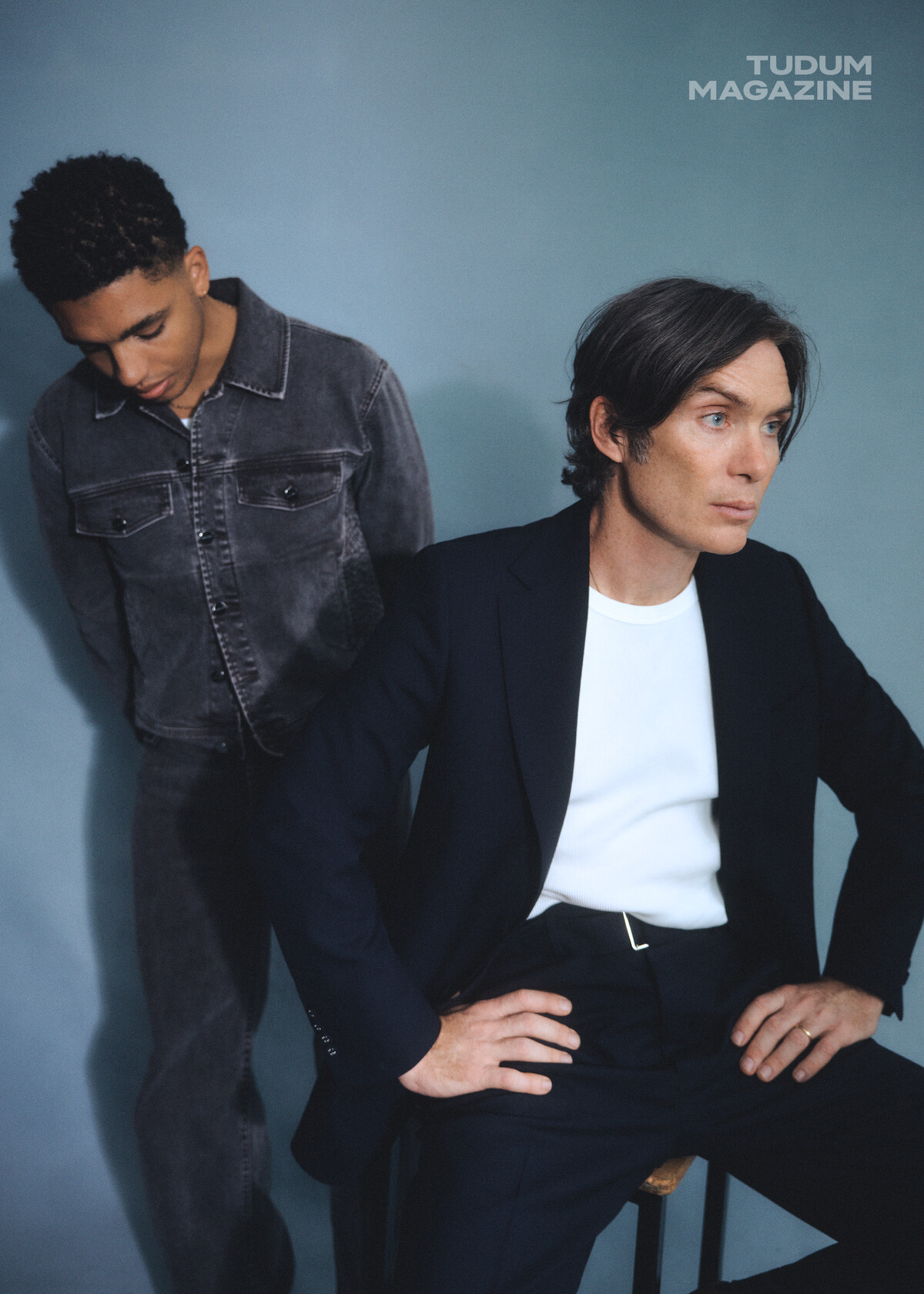 From left to right, Jay Lycurgo and Cillian Murphy pose together on set in front of a blue wall. Jay is standing in the background looking down and Cillian sits on a stool in the foreground looking to the distance.