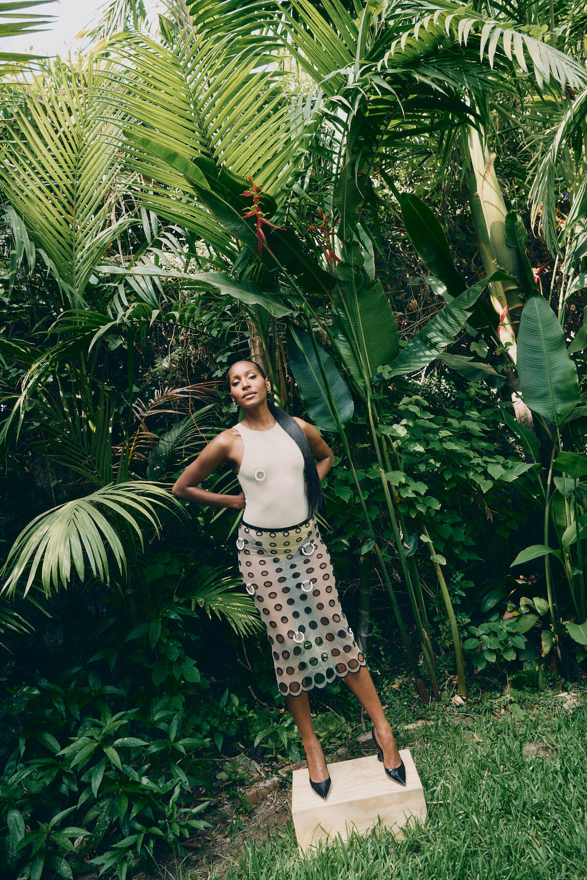 A woman poses outdoors in a lush, tropical garden, standing confidently on a stone surrounded by green plants and palm leaves, wearing a white top, patterned skirt, and black heels.