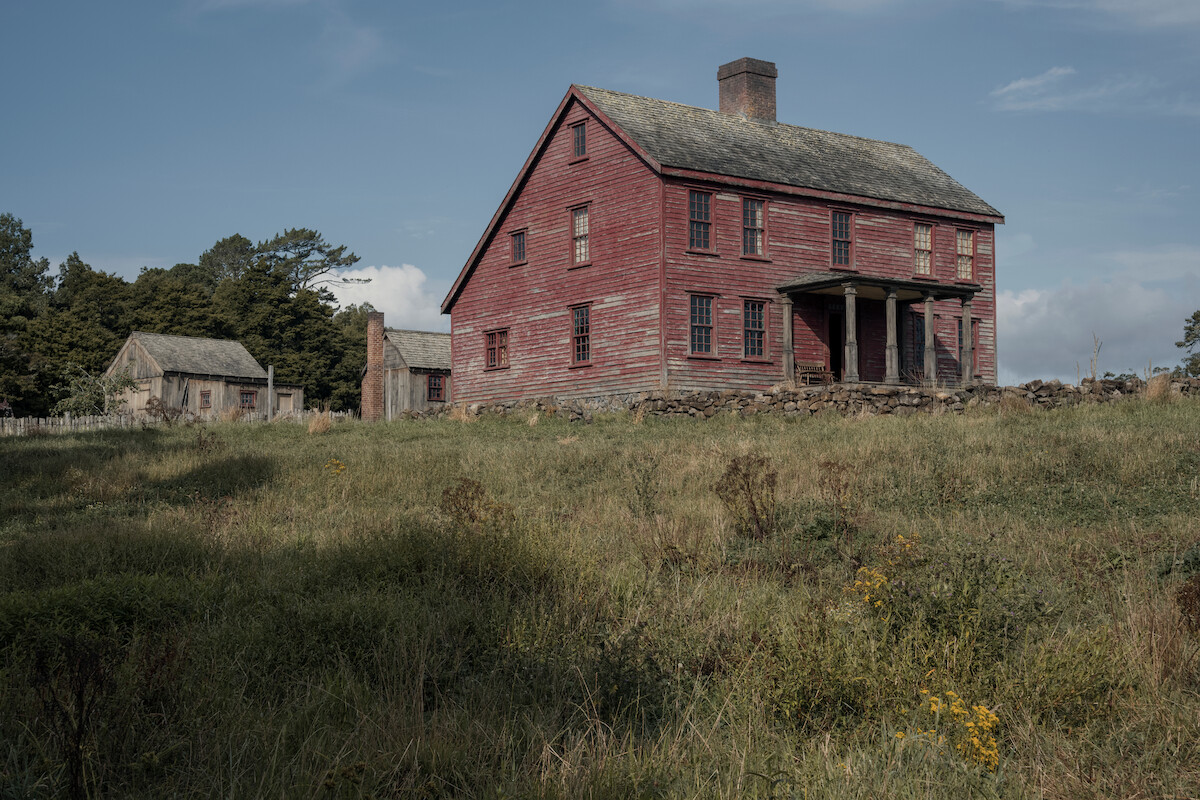 A weathered red wooden farmhouse with a front porch stands on a grassy hill, surrounded by overgrown fields, stone walls, and trees, with another rustic outbuilding in the background under a partly cloudy sky.