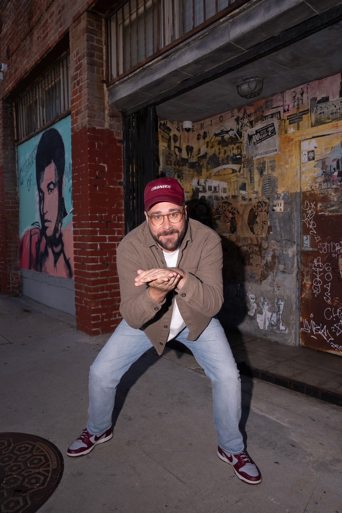 Man wearing red cap, glasses, brown jacket, and jeans squatting on urban sidewalk at night, posing with hands together, colorful mural and brick wall with graffiti in the background.