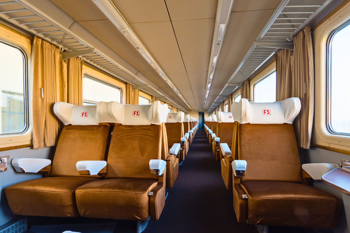 Empty vintage train carriage with brown cushioned seats, white headrests, wide windows with curtains, overhead luggage racks, and soft natural light creating a calm, comfortable, and inviting atmosphere.
