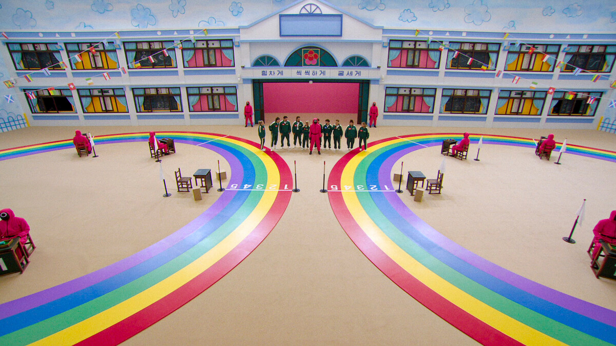 Players stand in a colorful, whimsical game arena with rainbow paths and costumed guards, set in a large, brightly decorated room resembling a school, creating a surreal and tense atmosphere.