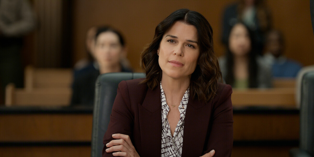 Woman in a blazer sits with folded arms in a courtroom, looking thoughtful; blurred audience members are seated behind her, suggesting a legal proceeding or trial setting.