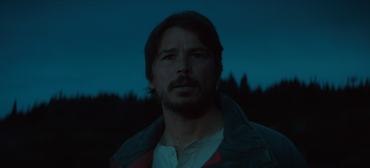 A man with facial hair stands outdoors at dusk, looking ahead with a concerned expression, silhouetted trees and a dark blue evening sky in the background.