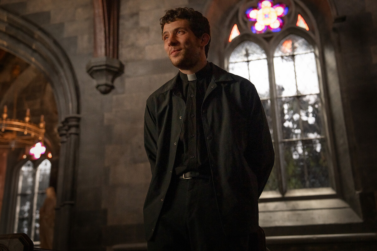 Young priest standing and smiling in a dimly lit, historic stone church with arched stained-glass windows, giving a warm and hopeful atmosphere.