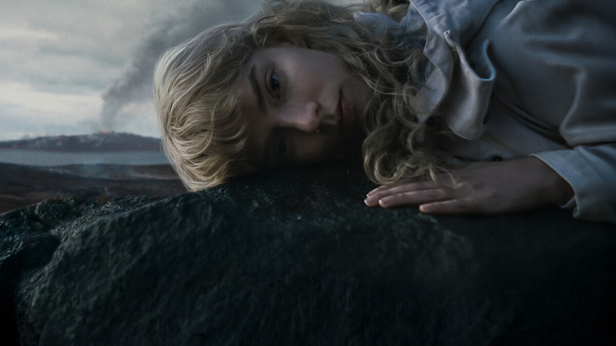 A woman with blonde curly hair lies on a dark rock outdoors, gazing somberly ahead. The background features a cloudy sky, distant mountains, and smoke rising, creating a dramatic and moody atmosphere.