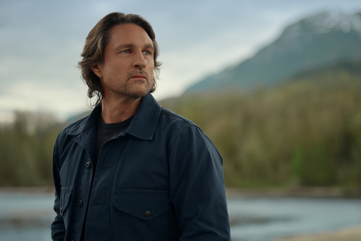 Man with medium-length hair wearing a dark jacket stands outdoors near a river, with a blurred forest and mountain in the background under a cloudy sky.