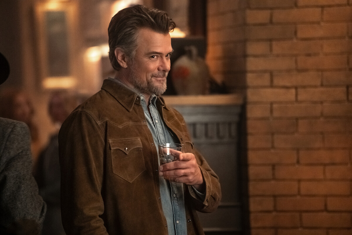 A man with gray hair and beard wearing a brown suede jacket stands indoors by a brick wall, holding a glass and smiling in a warmly lit, cozy setting that appears to be a social gathering or party.