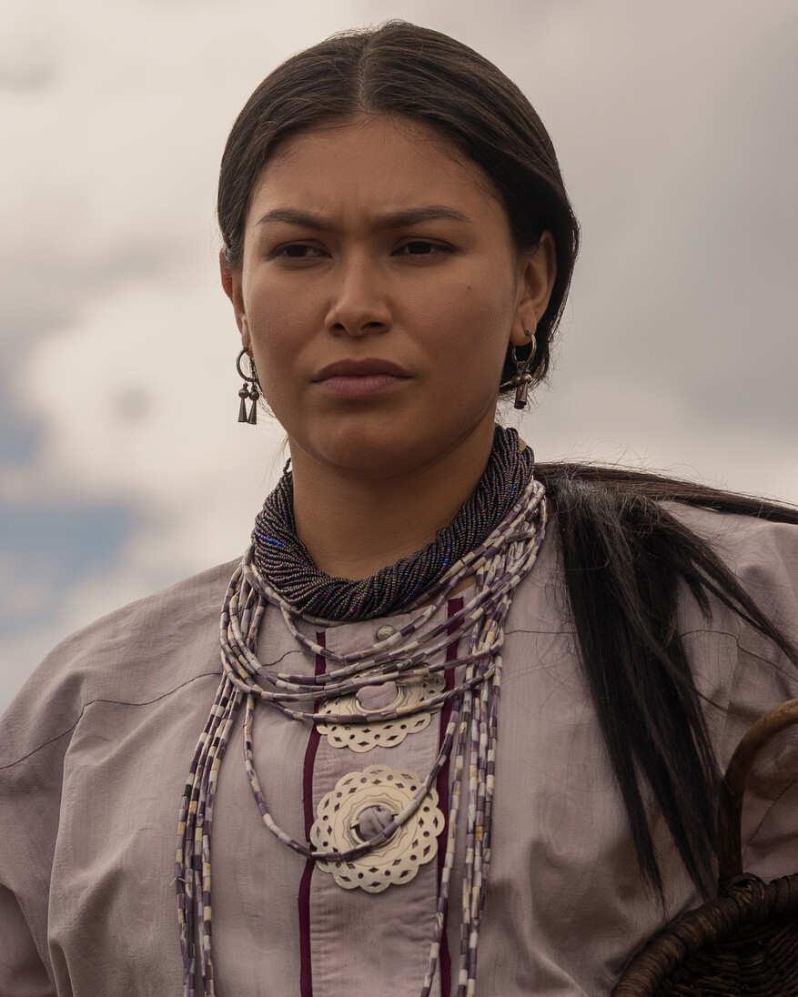 Young woman wearing traditional attire and layered beaded jewelry, standing outdoors with cloudy sky in background, looking serious and composed.