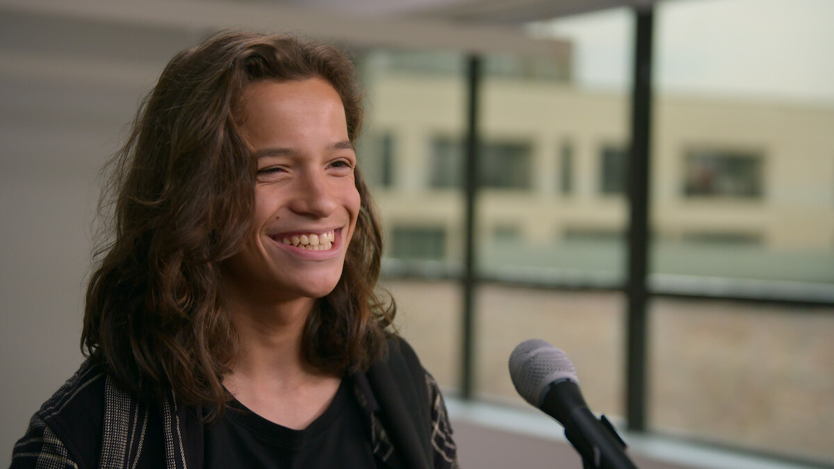 Smiling young person with long hair stands in front of a microphone indoors, with large windows and an urban background, giving a cheerful and relaxed mood.