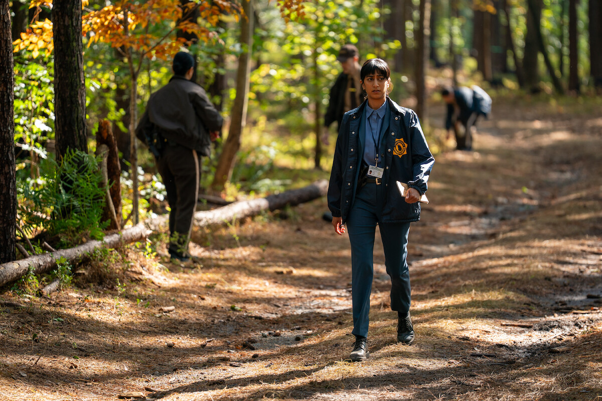 A woman in a detective jacket walks on a sunlit forest trail while officers search the woods in the background.