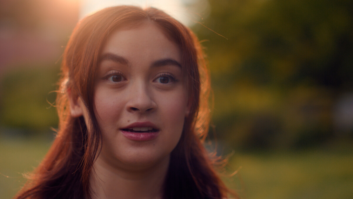 A young woman with long brown hair looks surprised outdoors in a park or garden setting during sunset, with soft natural lighting and a blurred green background.
