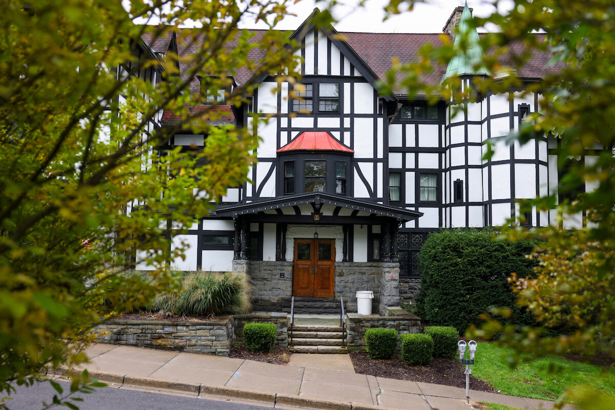 Tudor-style house with half-timbered exterior, large wooden doors, front steps, stone base, and red roof detail, surrounded by trees, bushes, grass, and sidewalk.