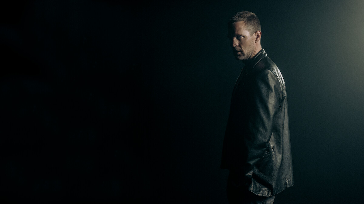 A man in a leather jacket stands in a dark, moody environment with dramatic lighting from behind, creating a tense and mysterious atmosphere.
