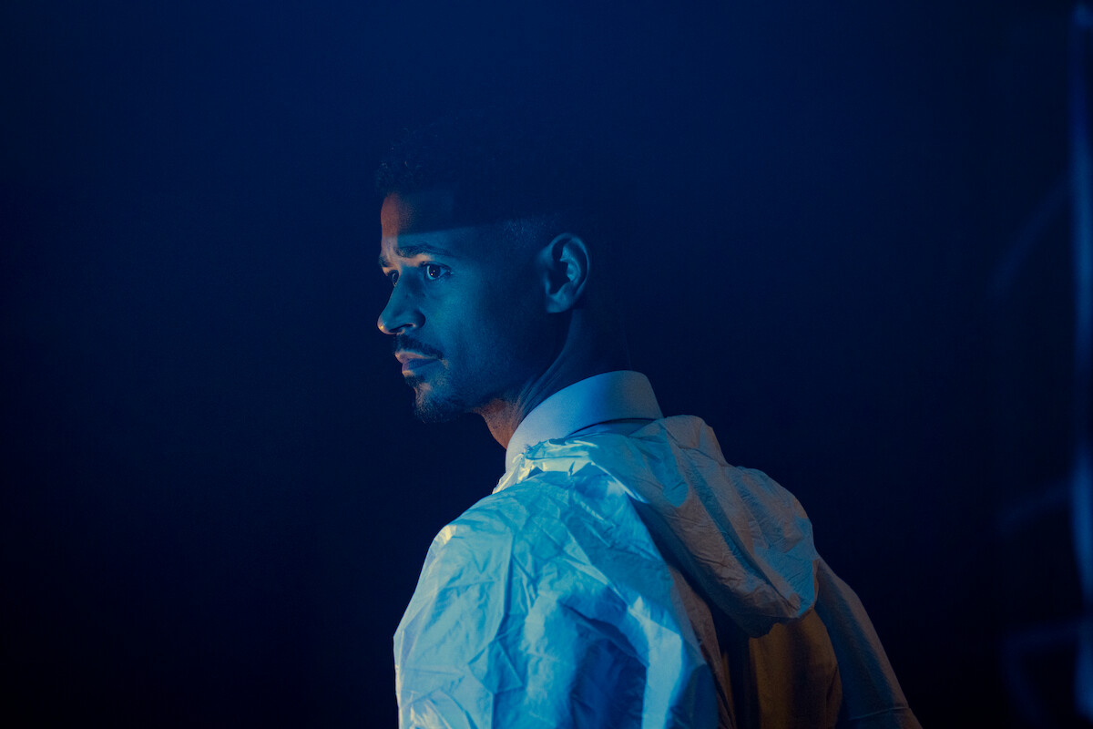 A man in a white jacket stands in a dimly lit, moody environment with blue lighting, looking serious as he gazes over his shoulder, evoking a suspenseful or contemplative mood.