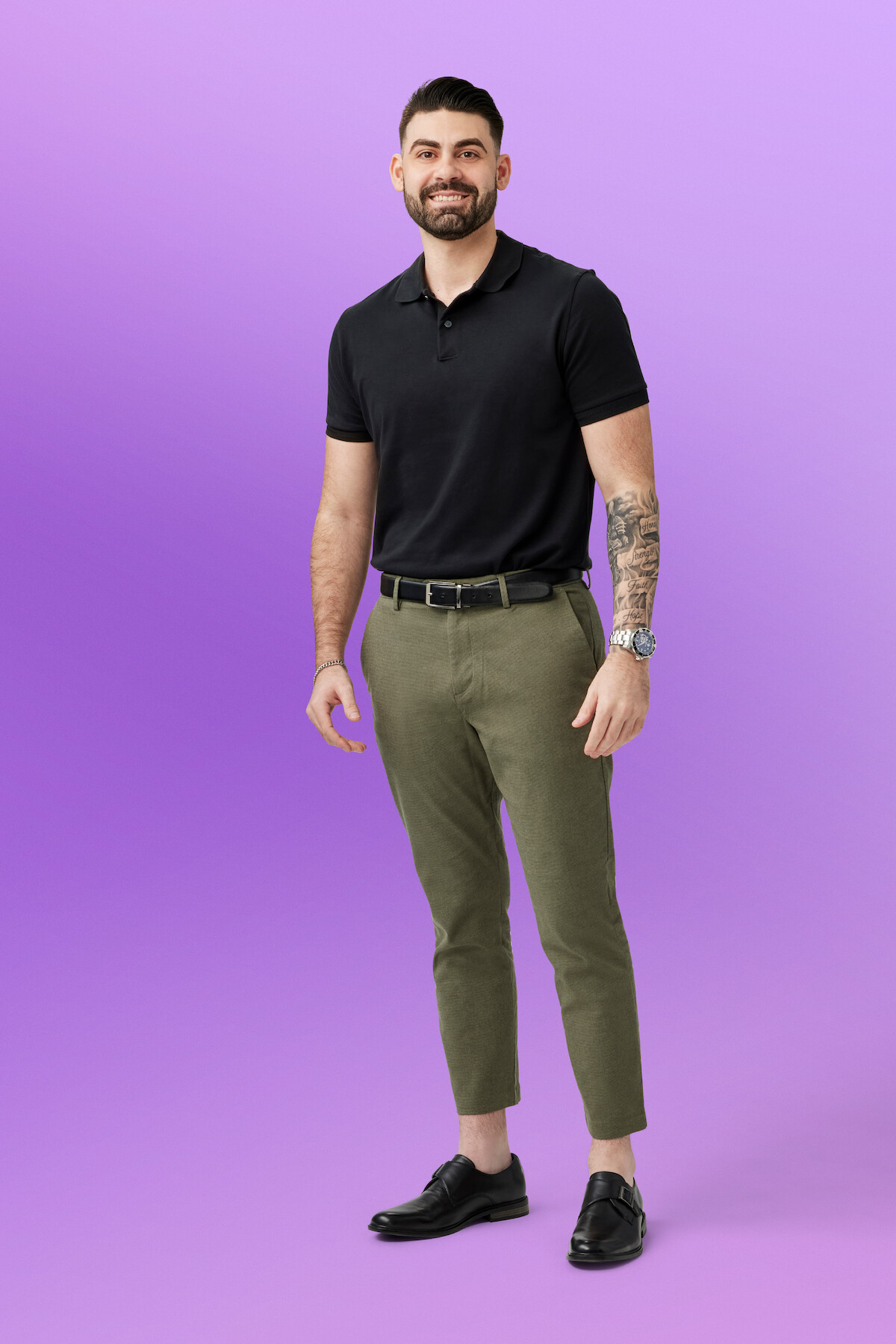 Mike in a black polo shirt, olive green pants, and black loafers standing and smiling in front of a plain purple background, in a well-lit studio setting.