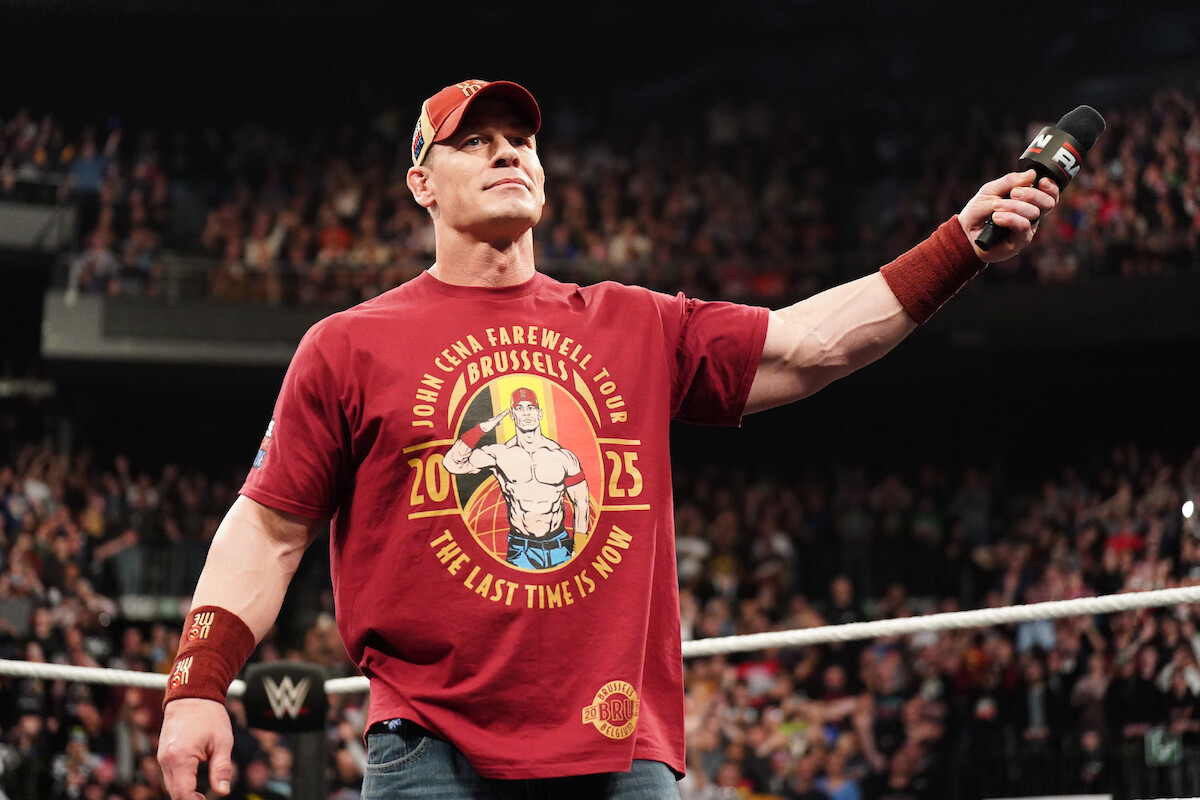 A muscular man in a red shirt and cap stands confidently in a wrestling ring holding a microphone, with a lively crowd in the background, creating an energetic and celebratory atmosphere at a professional wrestling event.