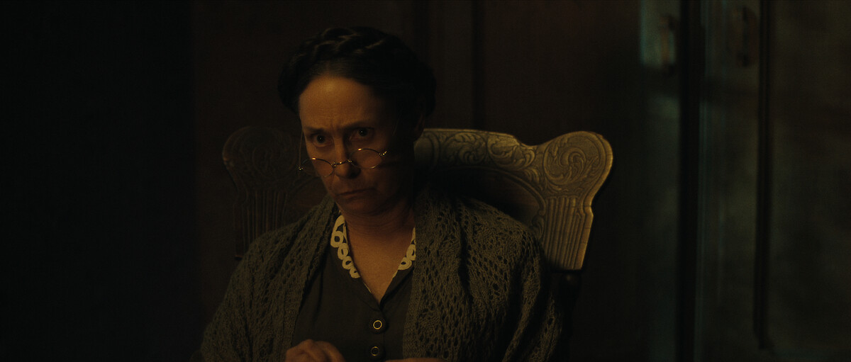 An older woman with glasses and braided hair sits in a carved wooden chair, looking sternly ahead in a dimly lit room.