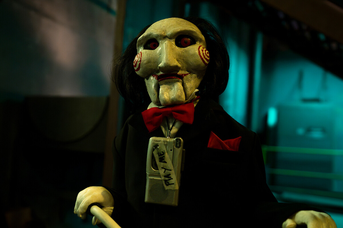 A creepy ventriloquist doll with white face, red eyes, and red bow tie stands in a dimly lit industrial room, with teal lighting and machinery in the background.