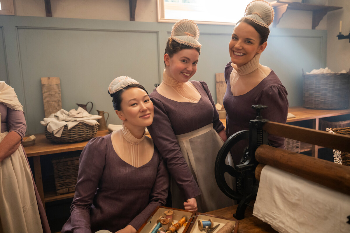 Três mulheres em roupas de época e toucas brancas posam sorrindo em uma lavanderia histórica, cercadas por cestos, roupas de cama e equipamentos antigos de lavar roupa, com luz natural suave entrando pela janela.