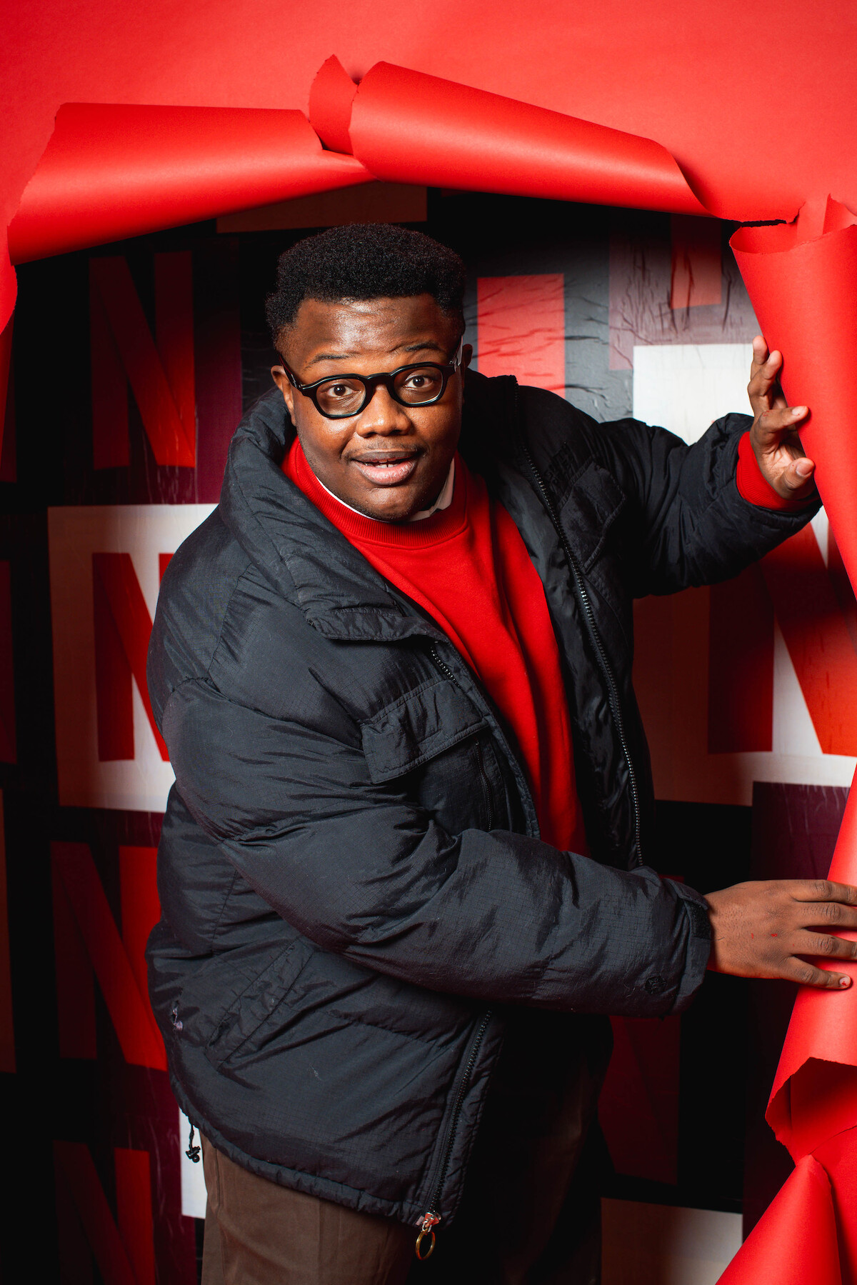 Um homem negro usando casaco e óculos se posiciona junto a um backdrop da Netflix e um buraco encenado na parede