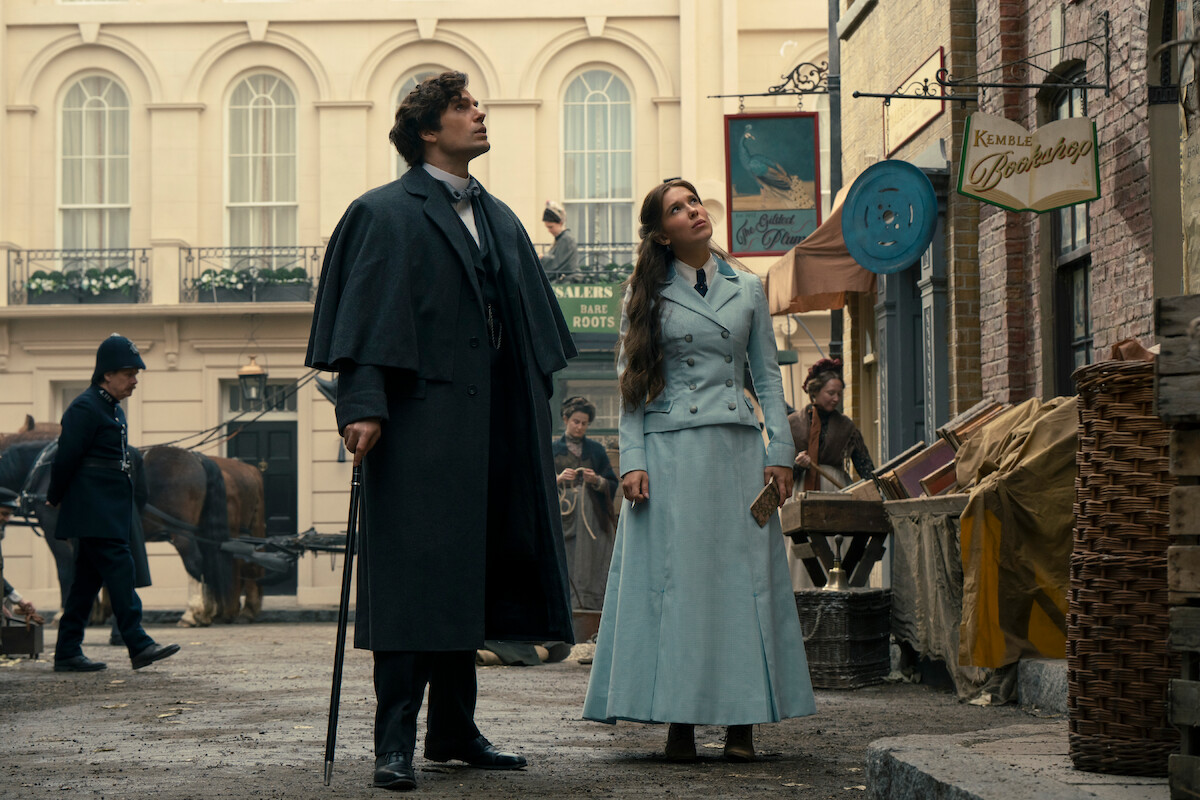 Two people in Victorian-era clothing stand on a cobblestone street, looking up, surrounded by market stalls and historical buildings; background includes horse-drawn carriage, pedestrians, and shop signs.
