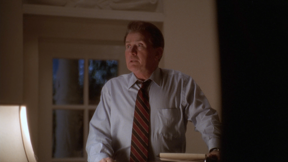 Man in dress shirt and tie stands by desk in softly lit office, looking upward with serious expression; window and lamp visible in background, nighttime outside.