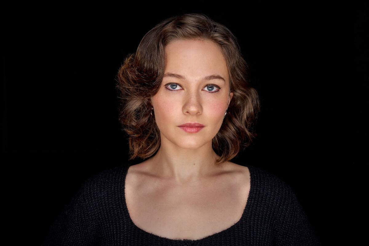 A young woman with short, wavy brown hair and blue eyes poses against a solid black background. She has a neutral, serene expression and wears a dark knit sweater, creating a calm and focused mood in a studio setting.