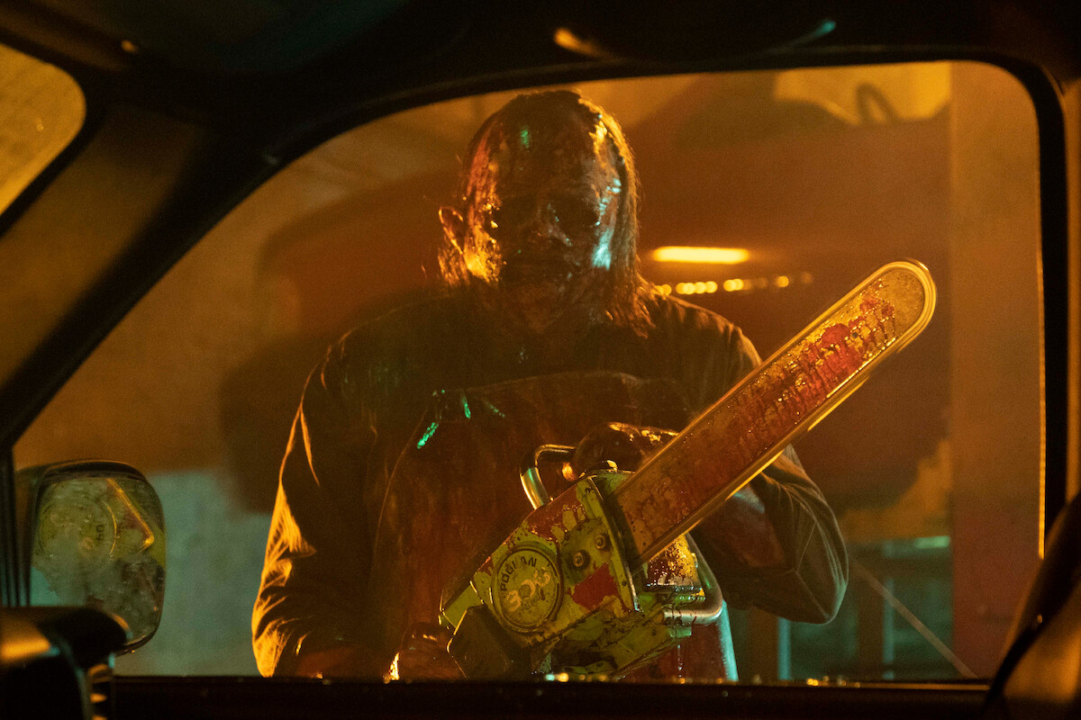 A menacing figure wearing a mask and apron stands outside a car window at night, holding a blood-stained chainsaw, in a dimly lit, suspenseful environment suggestive of a horror scene.