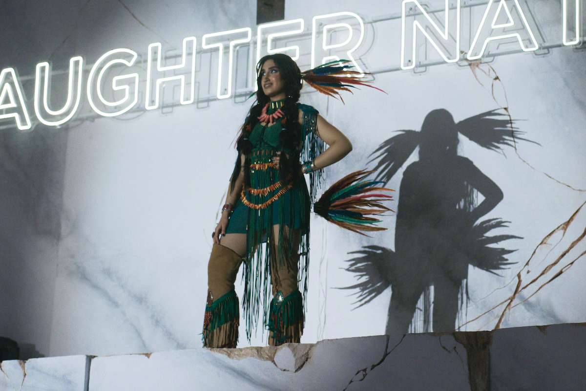 Woman in a feathered costume stands on a marble stage with cracked surfaces, illuminated by neon letters in the background, casting a dramatic shadow on the wall.