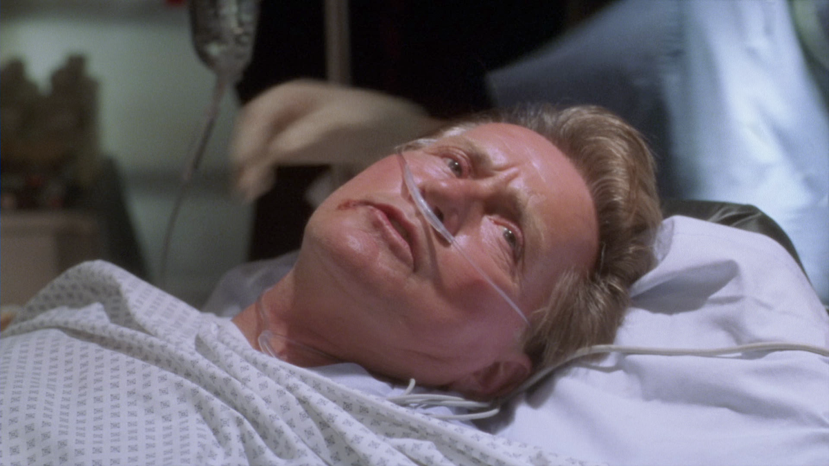 Elderly man lying in a hospital bed with an oxygen tube, looking distressed. Medical personnel and equipment are visible in a clinical setting, emphasizing a tense or serious medical situation.