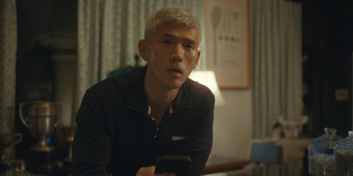 A man with short blond hair in a dark jacket sits indoors at a table, holding a phone. The setting is warmly lit with curtains, a lamp, trophies, and kitchen items visible in the background, creating a cozy domestic atmosphere.