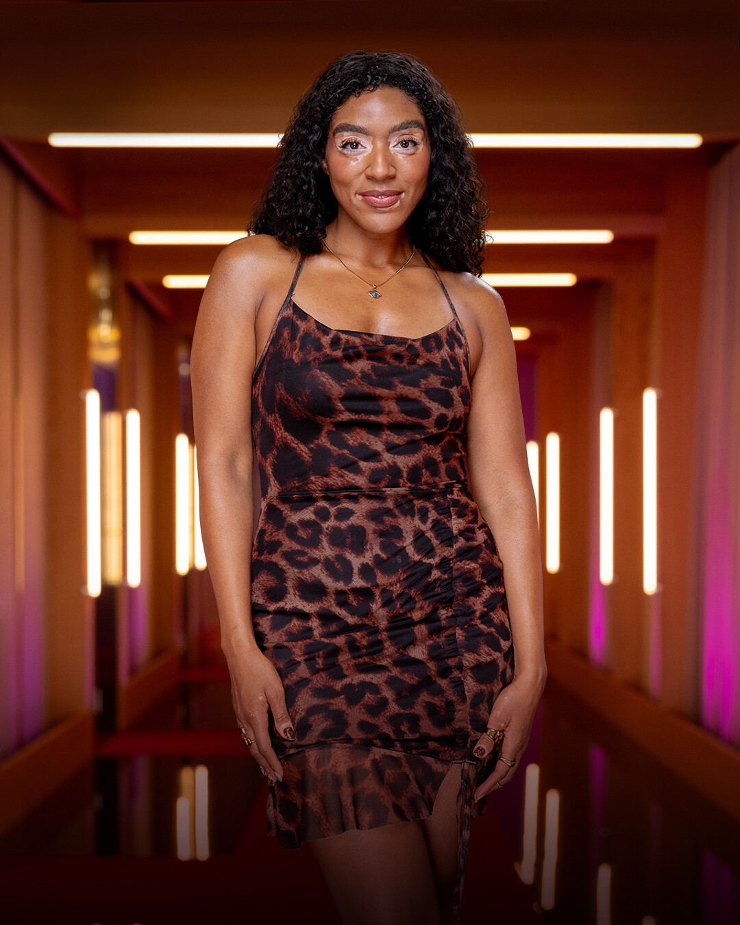 Woman with curly hair wearing a leopard print dress stands confidently in a modern hallway with warm lighting and vertical light fixtures, creating a stylish, contemporary atmosphere.