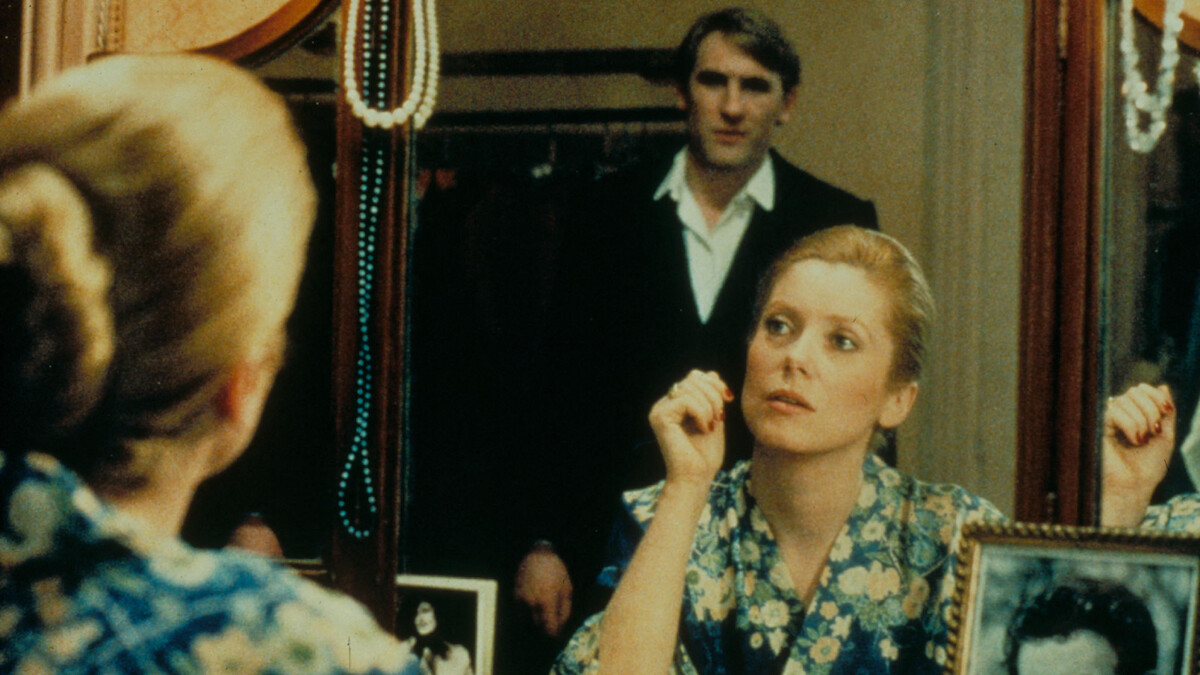 A woman in a floral robe sits at a dressing table, looking into a mirror with framed photos and jewelry. A man stands in the background, visible through the reflection, in a softly lit, intimate room.