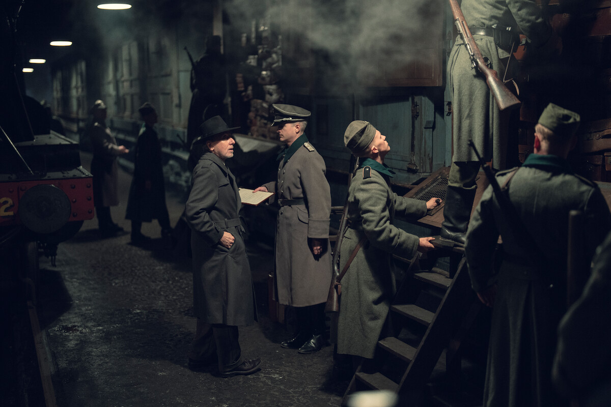 Soldiers in historical uniforms and a man in a hat stand beside a train in a dimly lit, smoke-filled railway station, checking papers and inspecting the train as others climb stairs into a boxcar.