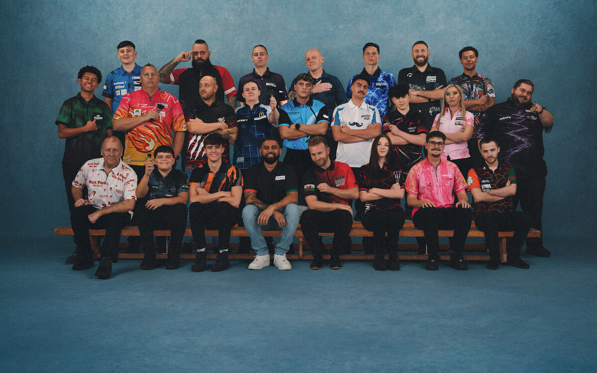 Large group of adults posing indoors against a blue backdrop. They wear colorful, patterned shirts and are seated and standing in three rows, smiling and making gestures, suggesting a team or group event is being commemorated.