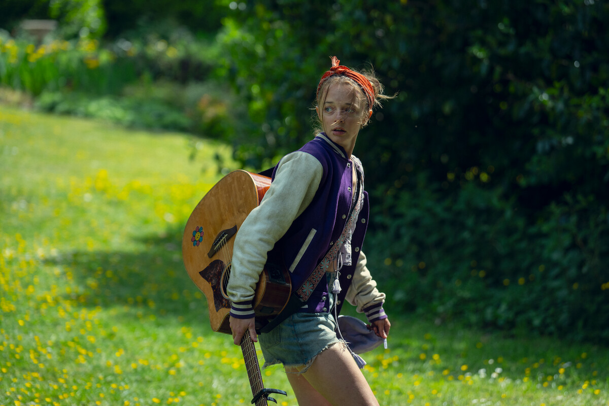 A young person in a varsity jacket and red headband holds an acoustic guitar outdoors in a grassy, sunlit field, looking alert and possibly concerned, with trees and flowers in the background.