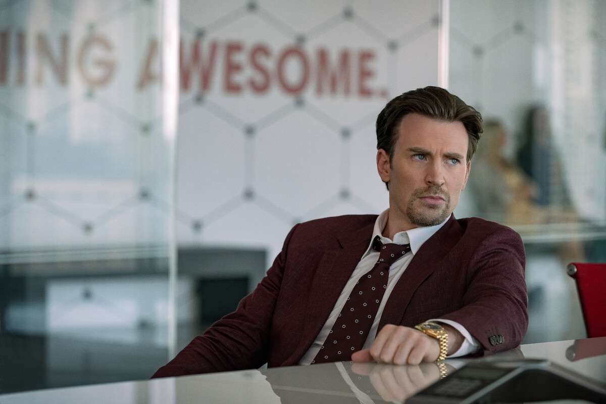 Chris Evans sits wearing a maroon suit and gold watch at a board room table in ‘Pain Hustlers.’