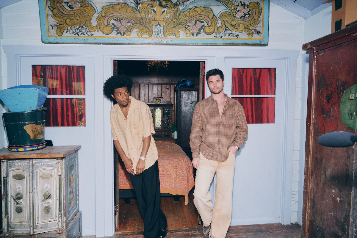 Two men standing in front of a rustic, ornately decorated room with blue wooden walls and red curtains, one leaning against the doorframe, vintage furniture and decorative elements visible around them.