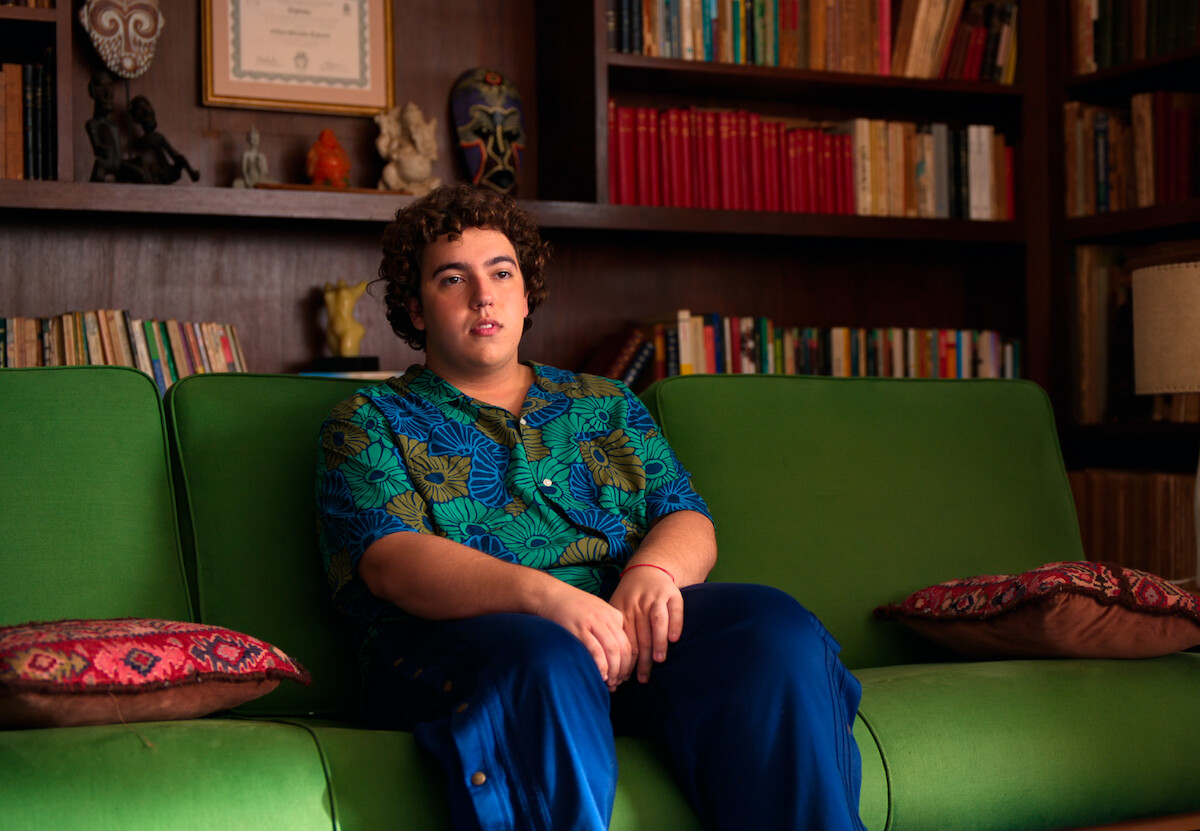 Homem jovem com blusa estampada em tons de verde e azul se senta em um sofá verde, numa sala de terapia