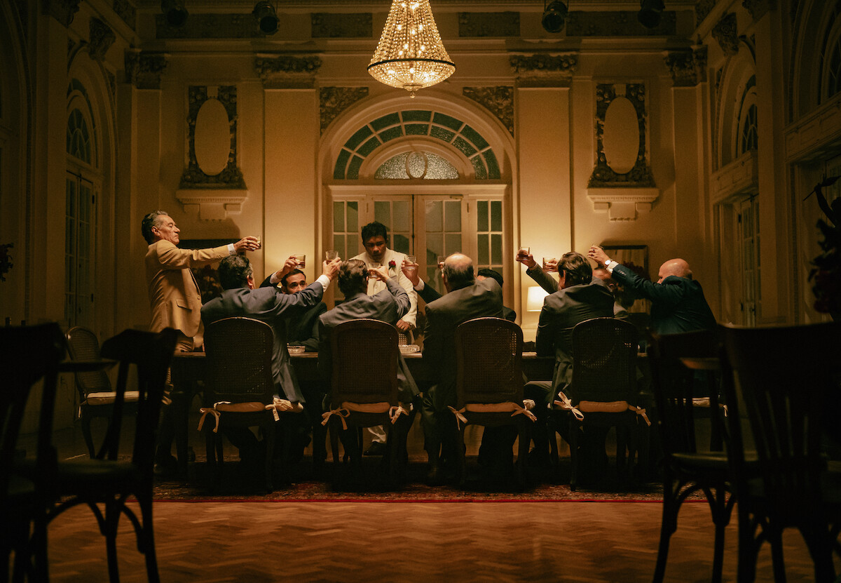 Homens em volta de uma mesa fazem um brinde em cena de “Os Donos do Jogoä