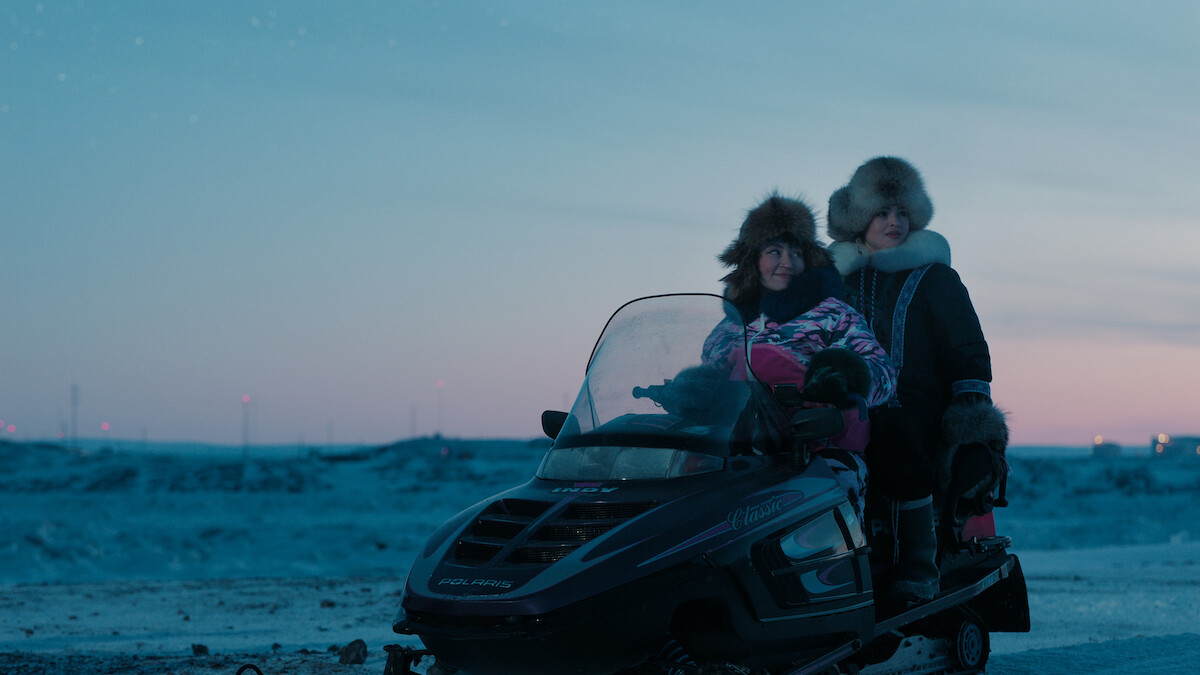 Two people in fur-lined winter clothing sit on a snowmobile in a snowy, icy landscape at dusk, with a softly lit sky and distant lights on the horizon.