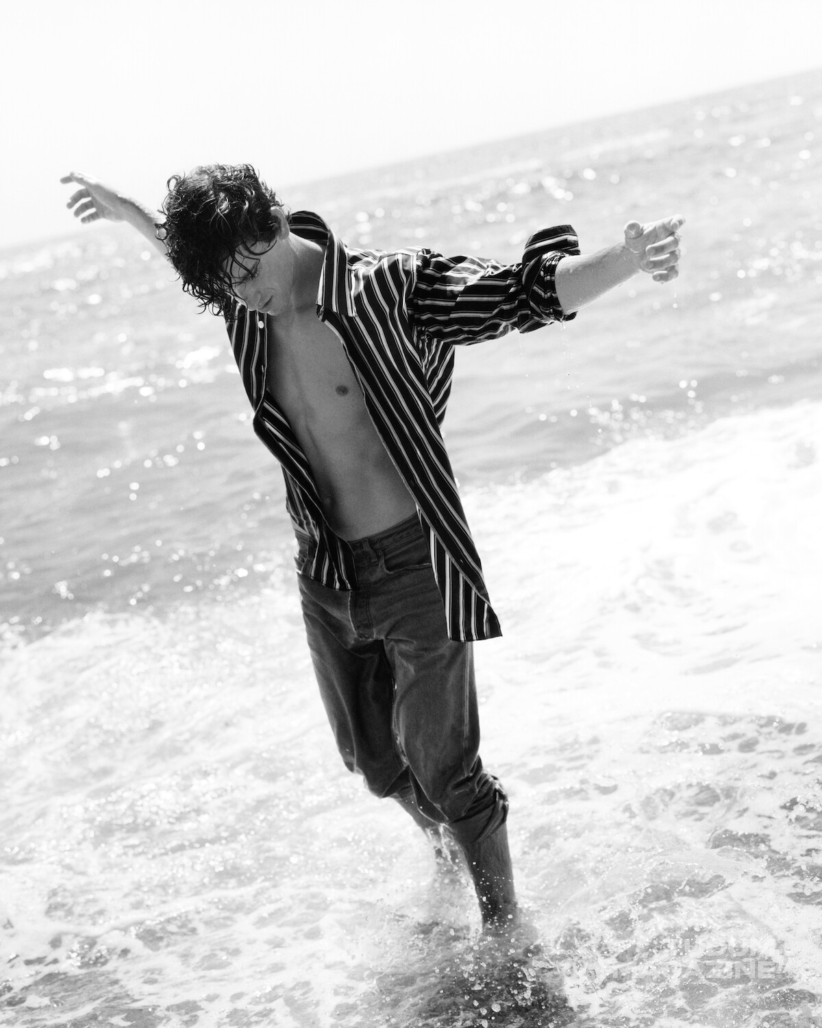 A person with wet hair and an open striped shirt stands in the ocean surf with arms outstretched, enjoying the beach. Black and white photo, waves around their legs, relaxed and carefree mood.