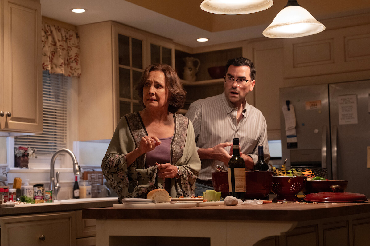 Two people stand in a warmly lit kitchen, looking surprised. Various food items, bottles, and kitchen utensils are on the counter. The environment is a home kitchen with beige cabinets and overhead lighting.