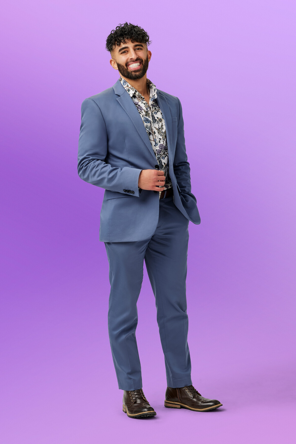 Haramol wearing a blue suit and floral shirt poses confidently while smiling in front of a solid purple background in a studio setting.
