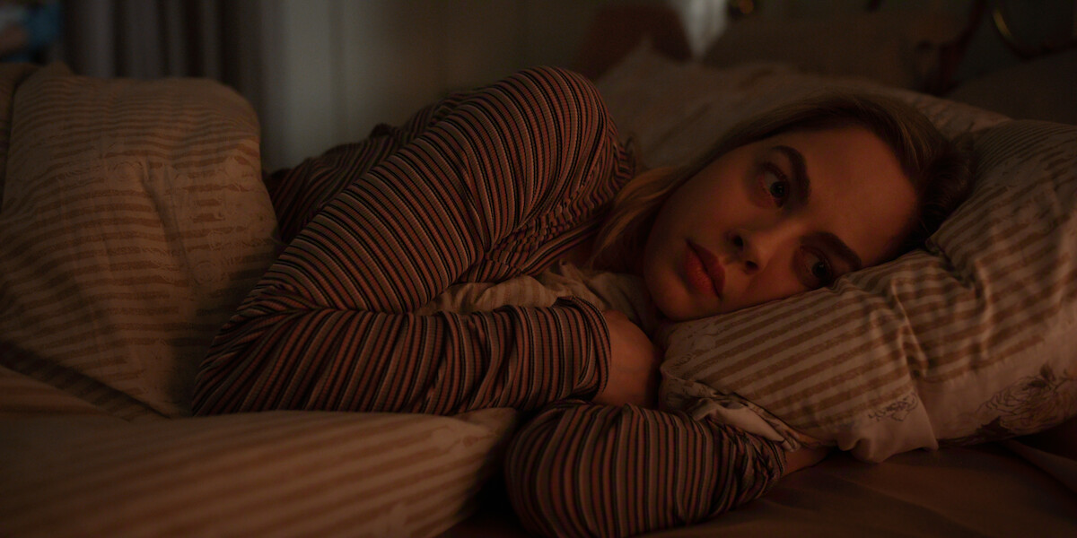Young woman lying in bed at night, wearing striped pajamas, clutching a pillow and staring thoughtfully, illuminated by soft, warm indoor lighting in a cozy bedroom setting.