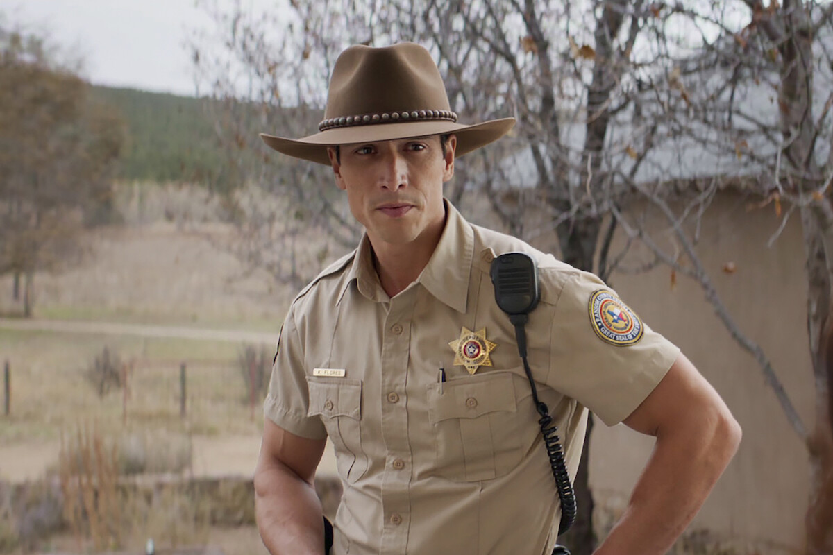 A law enforcement officer in a tan uniform and cowboy hat stands outdoors near a rural building with trees and open land in the background, looking directly at the camera.