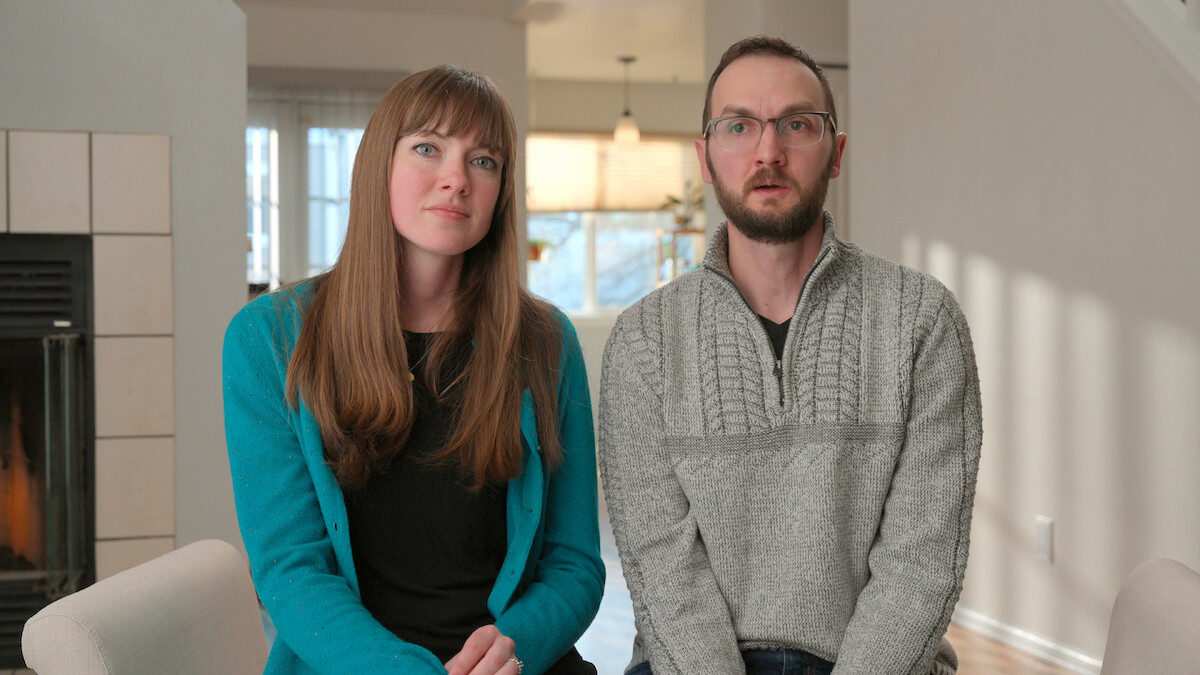 Two adults sitting indoors on chairs in a bright living room, with a fireplace on the left and natural light from large windows in the background.